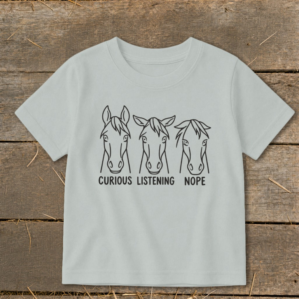 Light gray short-sleeved children's t-shirt featuring a black line art graphic of three horse heads with the text "CURIOUS," "LISTENING," and "NOPE."