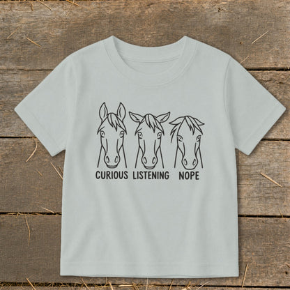 Light gray short-sleeved children's t-shirt featuring a black line art graphic of three horse heads with the text "CURIOUS," "LISTENING," and "NOPE."
