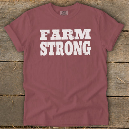 A mauve short-sleeved t-shirt with distressed white text reading FARM STRONG on a wooden background.