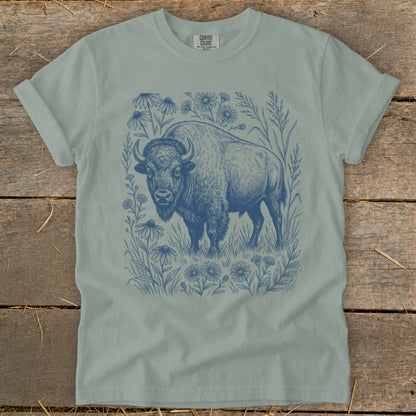 Light blue t-shirt with a dark blue linocut style print of a bison surrounded by wildflowers and grasses.