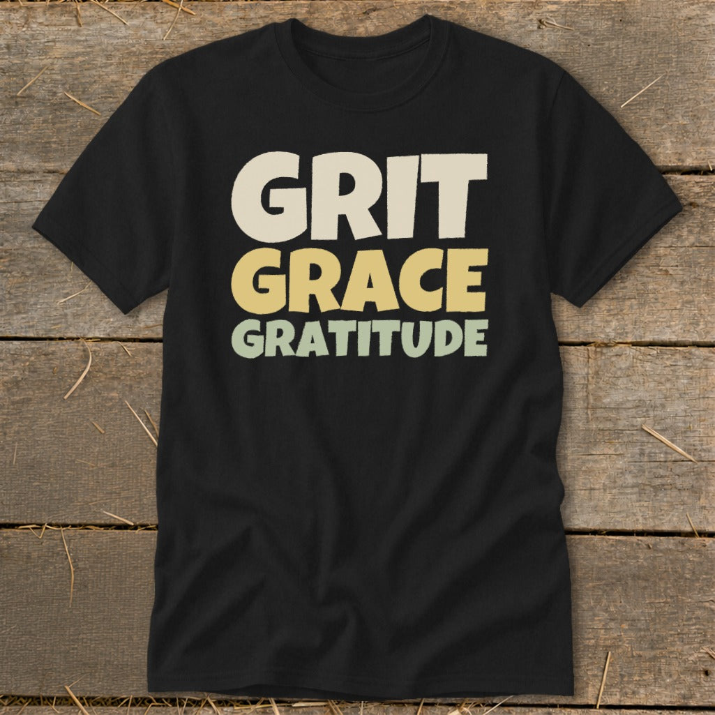 A black t-shirt printed with the stacked words Grit, Grace, and Gratitude in retro, earthy-toned block letters.