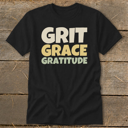 A black t-shirt printed with the stacked words Grit, Grace, and Gratitude in retro, earthy-toned block letters.