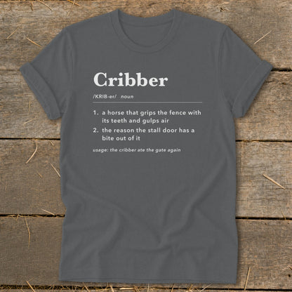 Dark grey t-shirt featuring dictionary-style text defining the word Cribber laid flat on wood planks.