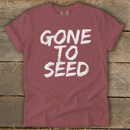 Maroon t-shirt with white text "GONE TO SEED" on a wooden background.