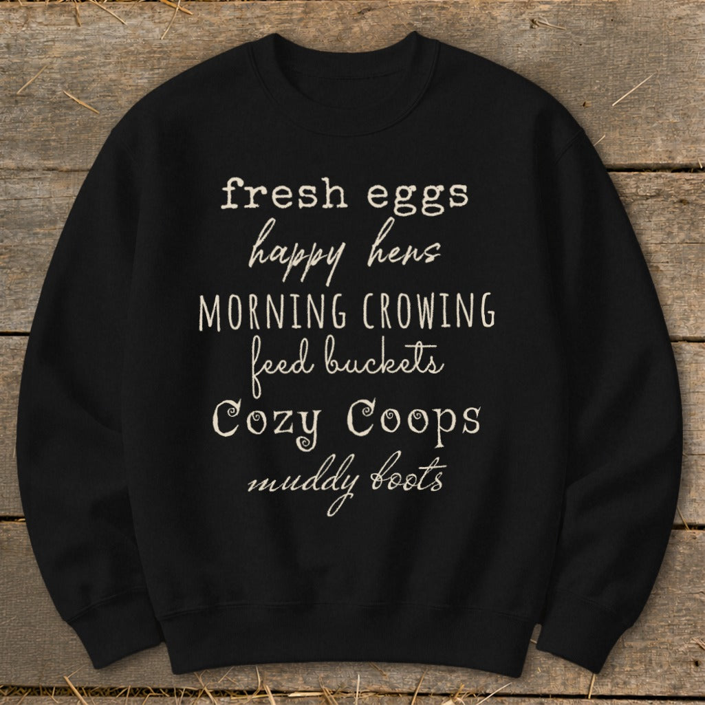 Black sweatshirt with white text listing "fresh eggs, happy hens, morning crowing, feed buckets, cozy coops, muddy boots".