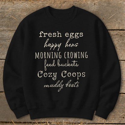 Black sweatshirt with white text listing "fresh eggs, happy hens, morning crowing, feed buckets, cozy coops, muddy boots".