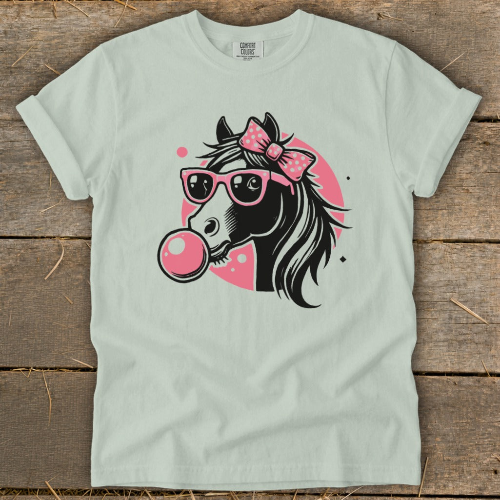 A light green t-shirt featuring a black horse wearing pink sunglasses, a dotted bow, and blowing a bubble.