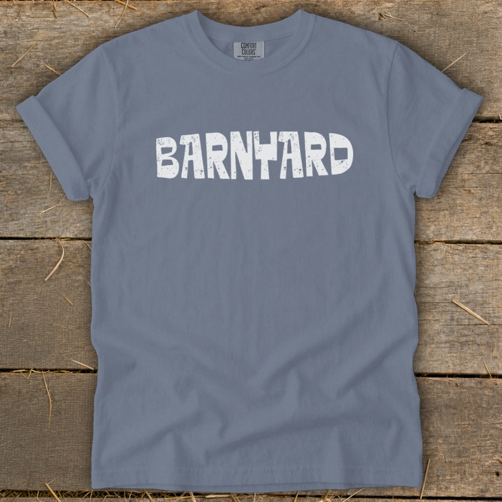 A slate blue crewneck t-shirt with rolled sleeves lies flat on a rustic wooden surface, featuring the word BARNYARD in distressed white text across the chest.