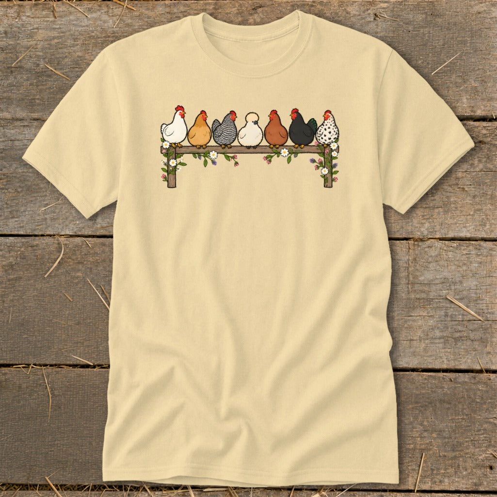A light yellow t-shirt features a graphic of seven colorful cartoon chickens perched on a flower-decorated wooden fence.