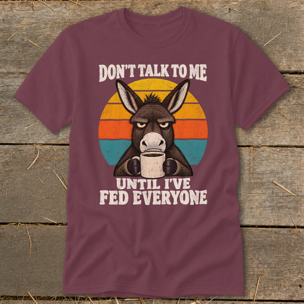 A maroon t-shirt featuring a grumpy donkey holding a coffee mug, overlaid on a sunset graphic with white text reading "Don't talk to me until I've fed everyone".
