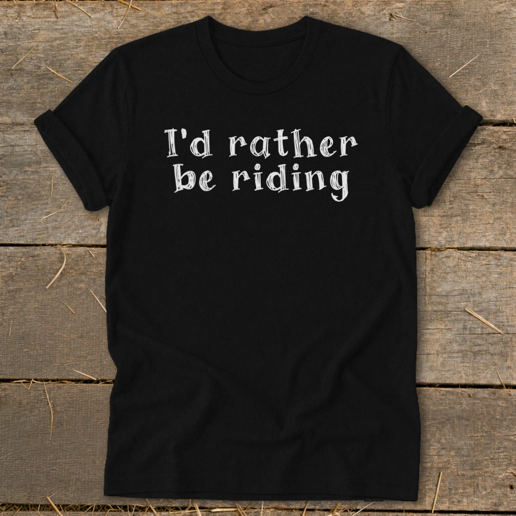 Black t-shirt with white text "I'd rather be riding" on a wooden background.