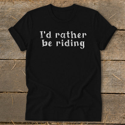 Black t-shirt with white text "I'd rather be riding" on a wooden background.