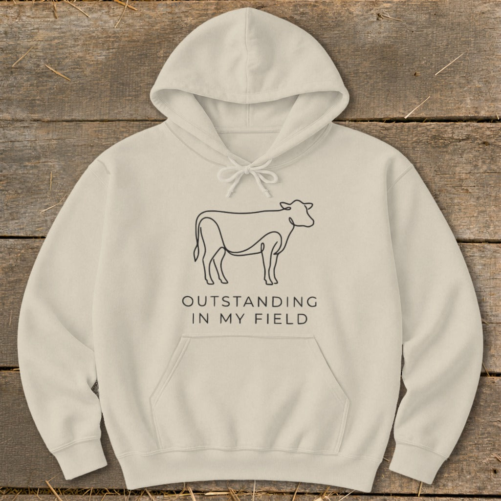 Cream hoodie with black line art cow and text "OUTSTANDING IN MY FIELD".