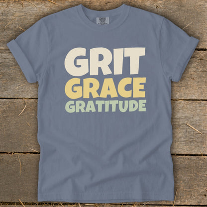 A blue crewneck t-shirt with rolled sleeves lies flat on a rustic wooden surface, featuring the words Grit Grace Gratitude in multi-colored, block-style lettering.