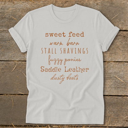 White t-shirt with brown text: sweet feed, warm barn, stall shavings, fuzzy ponies, saddle leather, dusty boots.