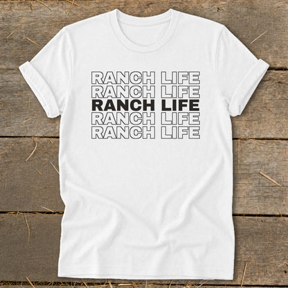 A white crewneck t-shirt with rolled sleeves lies flat on a rustic wooden surface, featuring centered black text reading RANCH LIFE repeated in five stacked rows, with the middle row solid black.