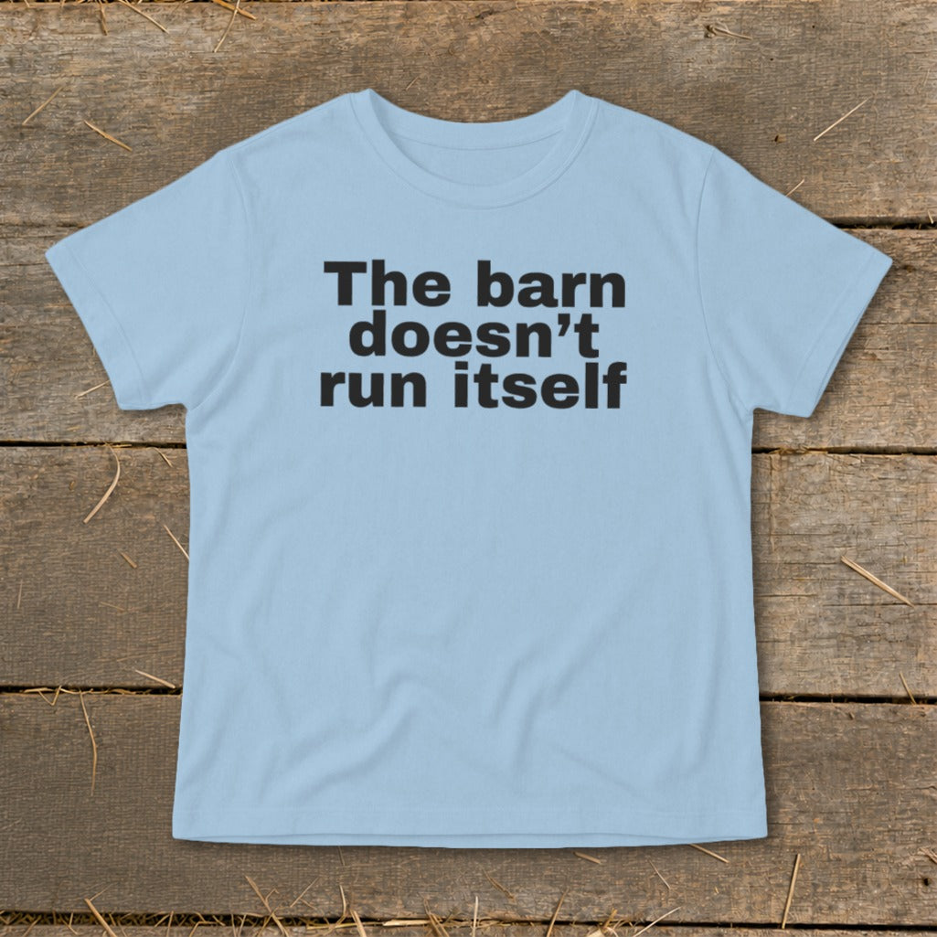 Light blue t-shirt with black text "The barn doesn't run itself" on a wooden background.