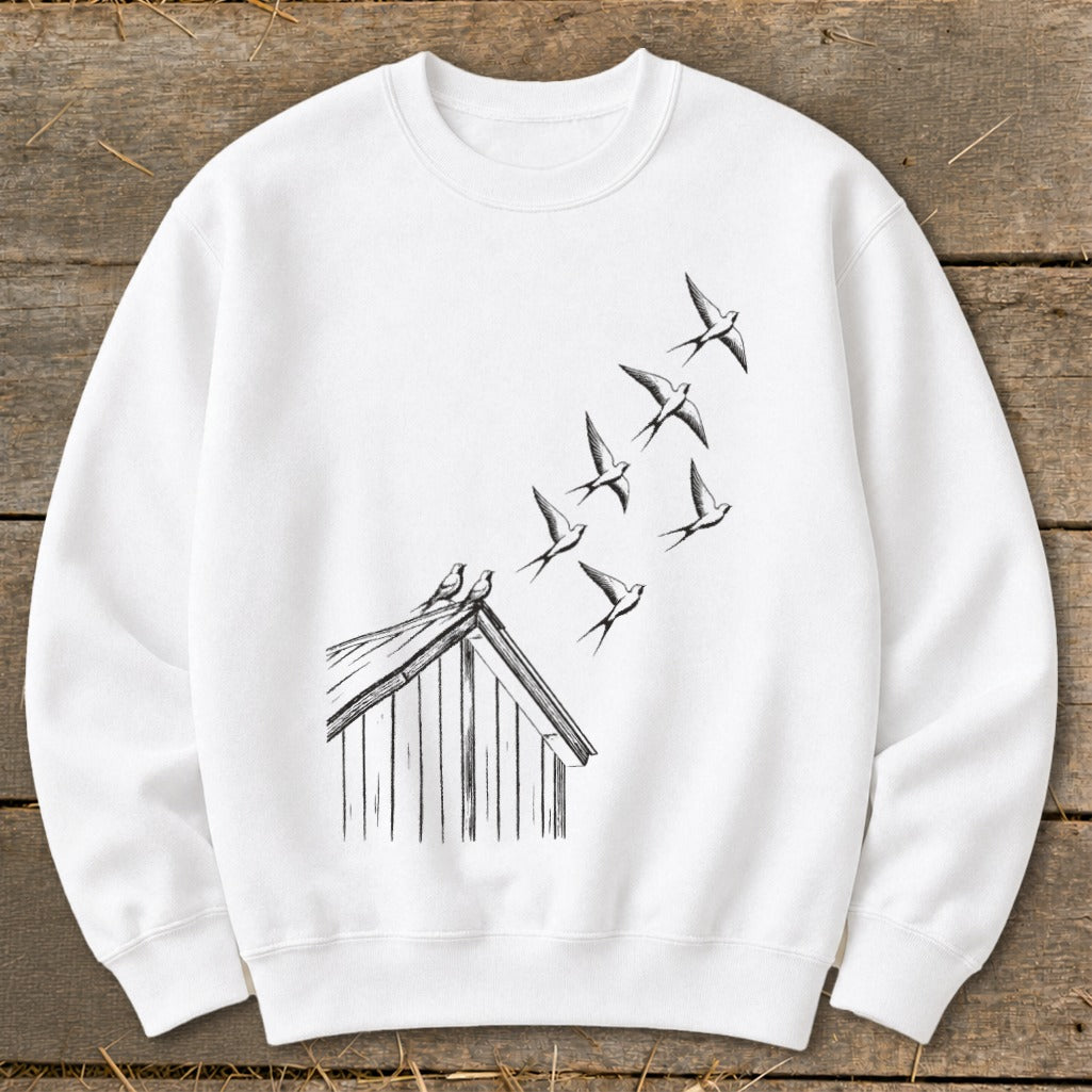 A white crewneck sweatshirt featuring a black sketch of birds flying from a small wooden shed.