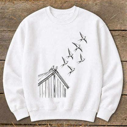 A white crewneck sweatshirt featuring a black sketch of birds flying from a small wooden shed.