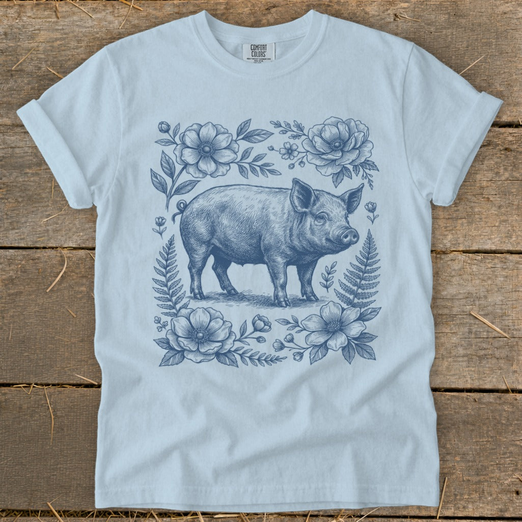 Light blue t-shirt with a blue pig illustration surrounded by flowers and ferns.