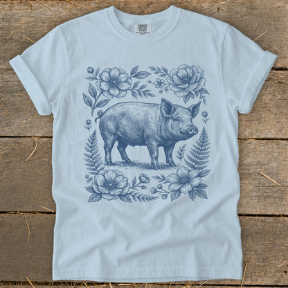 Light blue t-shirt with a blue pig illustration surrounded by flowers and ferns.