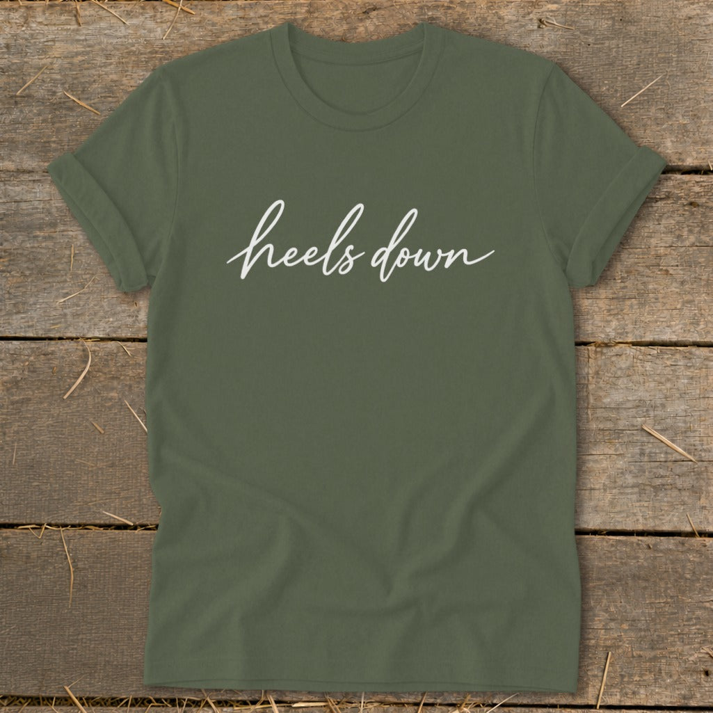 Olive green t-shirt with white cursive text "heels down" on a wooden background.