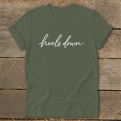 Olive green t-shirt with white cursive text "heels down" on a wooden background.