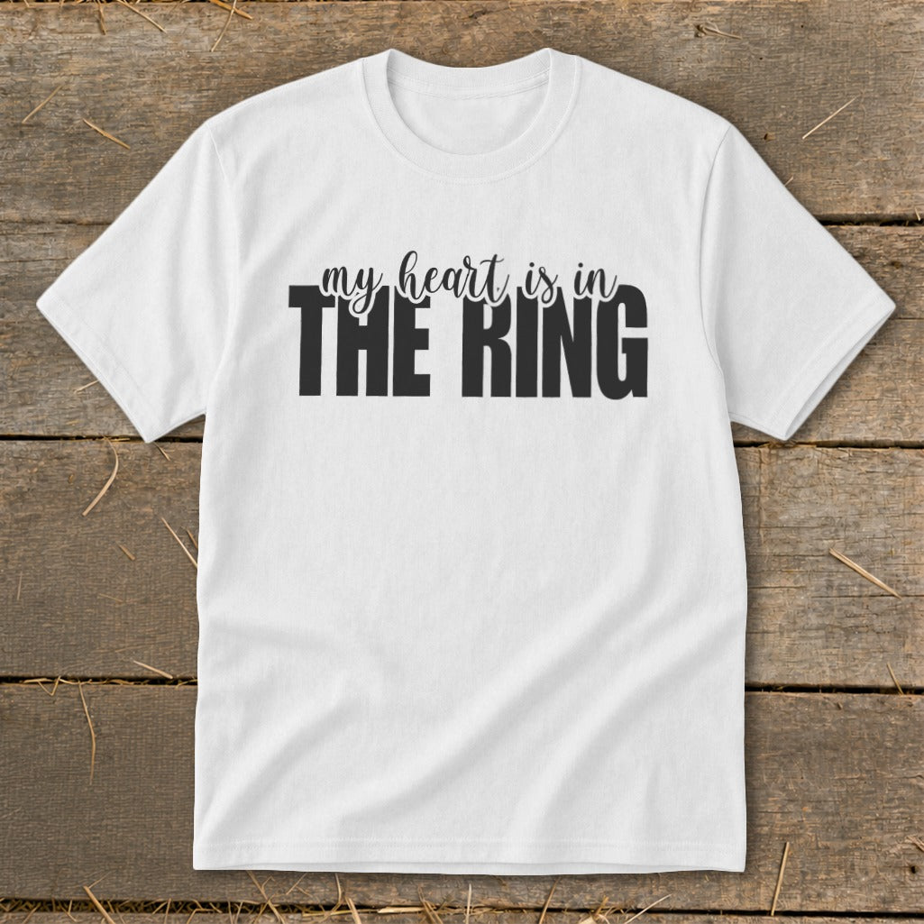 A white short-sleeved t-shirt with black text that reads "my heart is in THE RING" lies flat on a rustic wooden background.