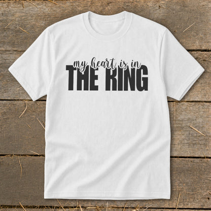 A white short-sleeved t-shirt with black text that reads "my heart is in THE RING" lies flat on a rustic wooden background.
