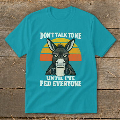A teal t-shirt featuring a graphic of a grumpy donkey drinking coffee and text reading, "DON'T TALK TO ME UNTIL I'VE FED EVERYONE."