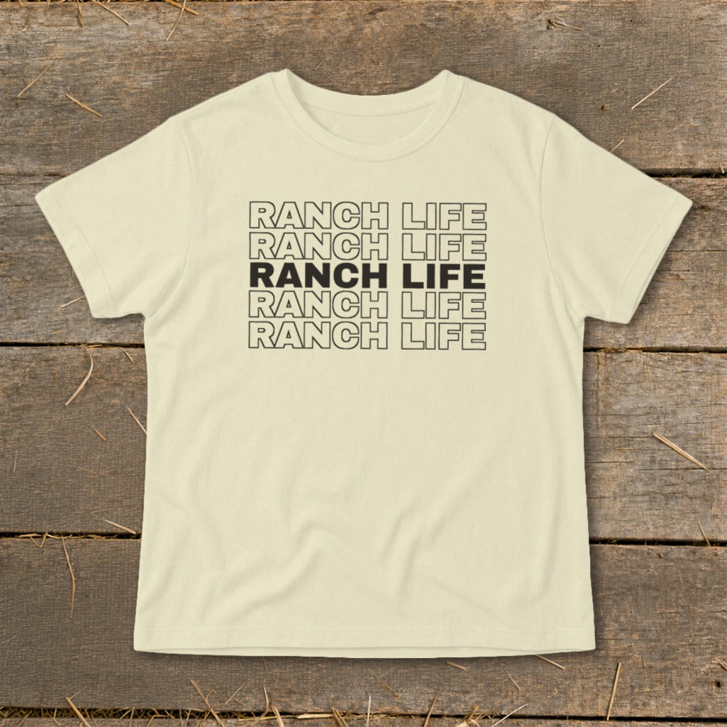 A cream-colored short-sleeved t-shirt featuring the text Ranch Life repeated in a stacked block design on a rustic wooden background.