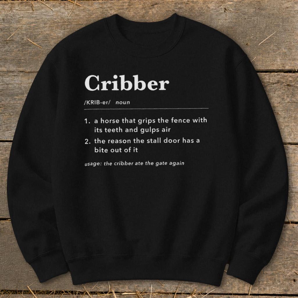 A black crewneck sweatshirt printed with white dictionary-style text defining the word Cribber laid on a wooden surface.