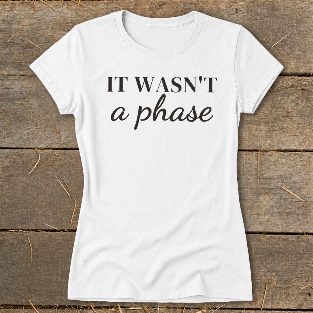 White t-shirt with black text reading "IT WASN'T a phase" on a wooden background.