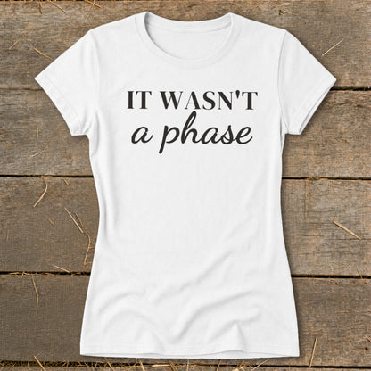 White t-shirt with black text reading "IT WASN'T a phase" on a wooden background.