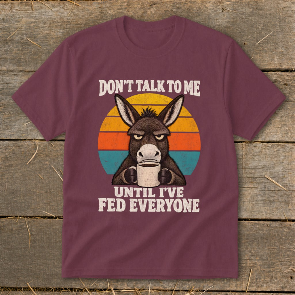 Maroon t-shirt featuring a grumpy donkey holding a mug against a striped sunset graphic with text.