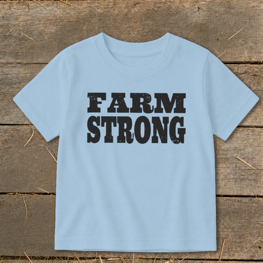A light blue children's t-shirt printed with the text FARM STRONG in black block letters, laid flat on a rustic wooden background.