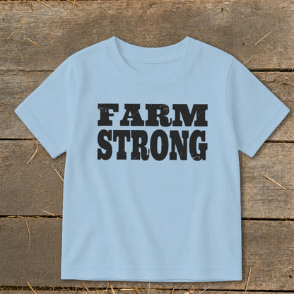 A light blue children's t-shirt printed with the text FARM STRONG in black block letters, laid flat on a rustic wooden background.