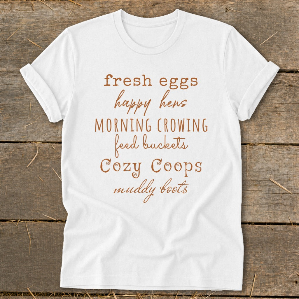White t-shirt with brown text listing "fresh eggs, happy hens, MORNING CROWING, feed buckets, Cozy Coops, muddy boots".