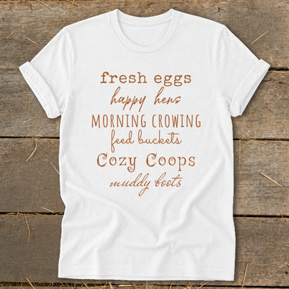 White t-shirt with brown text listing "fresh eggs, happy hens, MORNING CROWING, feed buckets, Cozy Coops, muddy boots".