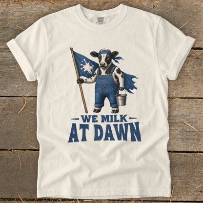White t-shirt with a cow wearing overalls, cape, and holding a milk splash flag and bucket. Text reads "WE MILK AT DAWN."