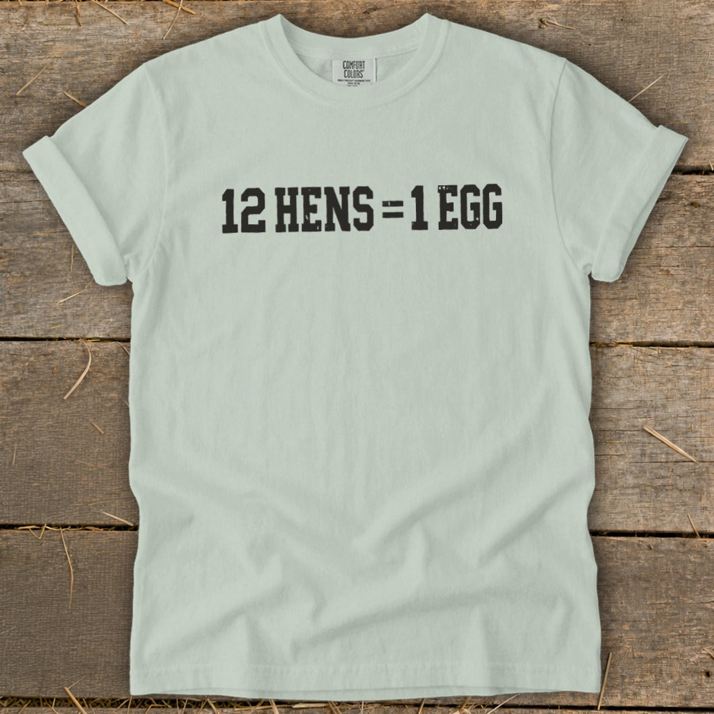 Light green t-shirt with rolled sleeves featuring the black distressed text 12 HENS = 1 EGG laid flat on a rustic wooden background.