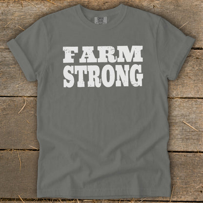 A grey t-shirt featuring the distressed white text "FARM STRONG" laid flat on a wooden surface.