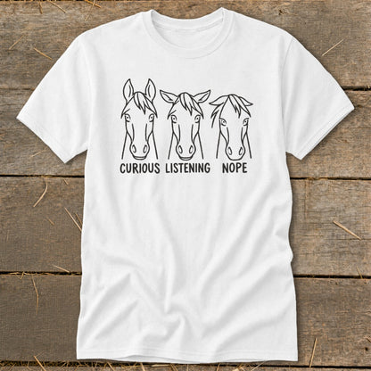 A white t-shirt featuring a black line drawing of three horse heads with the captions "Curious," "Listening," and "Nope" displayed on a wooden background.