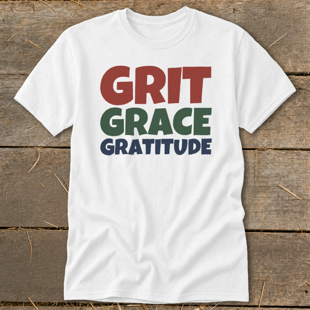 A white crewneck t-shirt featuring stacked text that reads "GRIT," "GRACE," and "GRATITUDE" in red, green, and blue.