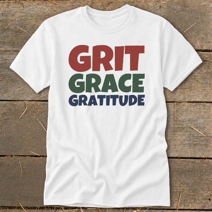 A white crewneck t-shirt featuring stacked text that reads "GRIT," "GRACE," and "GRATITUDE" in red, green, and blue.