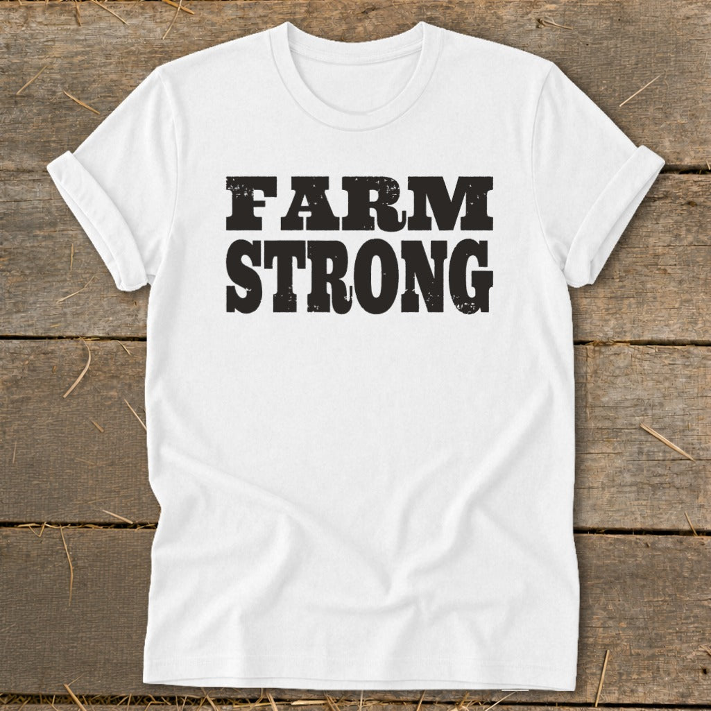 A white short-sleeved t-shirt with black distressed lettering reading FARM STRONG lies flat on a wooden surface.