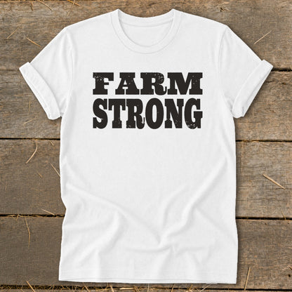 A white short-sleeved t-shirt with black distressed lettering reading FARM STRONG lies flat on a wooden surface.