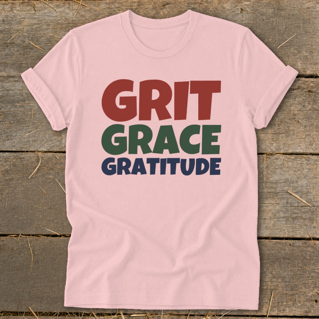 A pink crewneck t-shirt with short sleeves displays stacked text reading GRIT GRACE GRATITUDE.