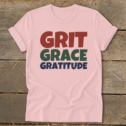 A pink crewneck t-shirt with short sleeves displays stacked text reading GRIT GRACE GRATITUDE.