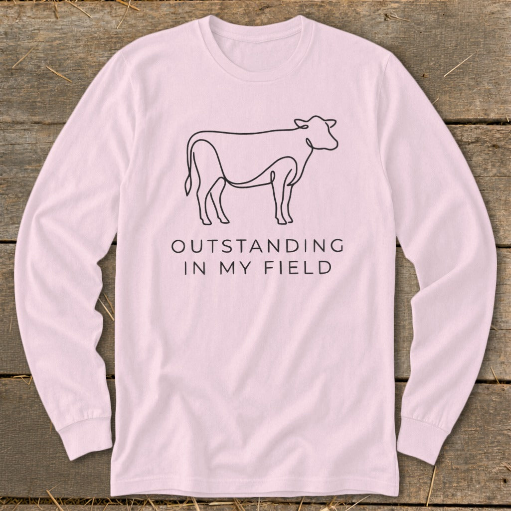 Pink long-sleeve shirt with cow line art and text "OUTSTANDING IN MY FIELD".